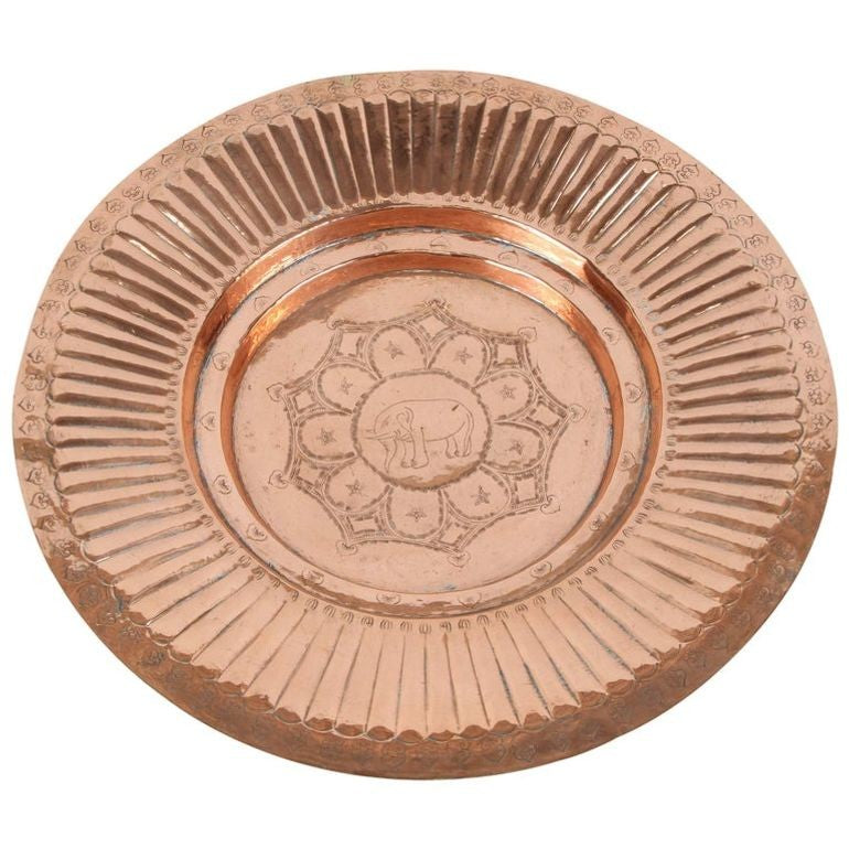 A polished round copper tray with a geometric design in the middle and an elephant in the center, against a light background.