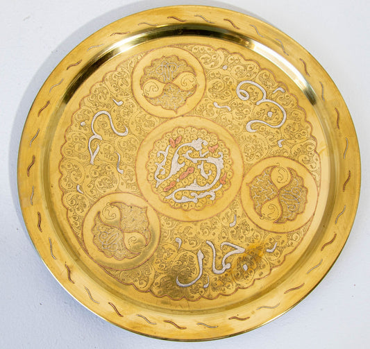 A round gold-colored brass tray with geometric patterns and text inlay, possibly used for serving or as a decorative piece.