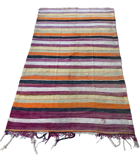 A vintage Moroccan tribal rug with a striped pattern in various colors including orange, purple, and black, showing signs of wear and distress.