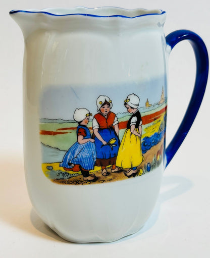 White pitcher with blue rim and handle featuring a colorful illustration of three women in traditional Dutch clothing on a white background.