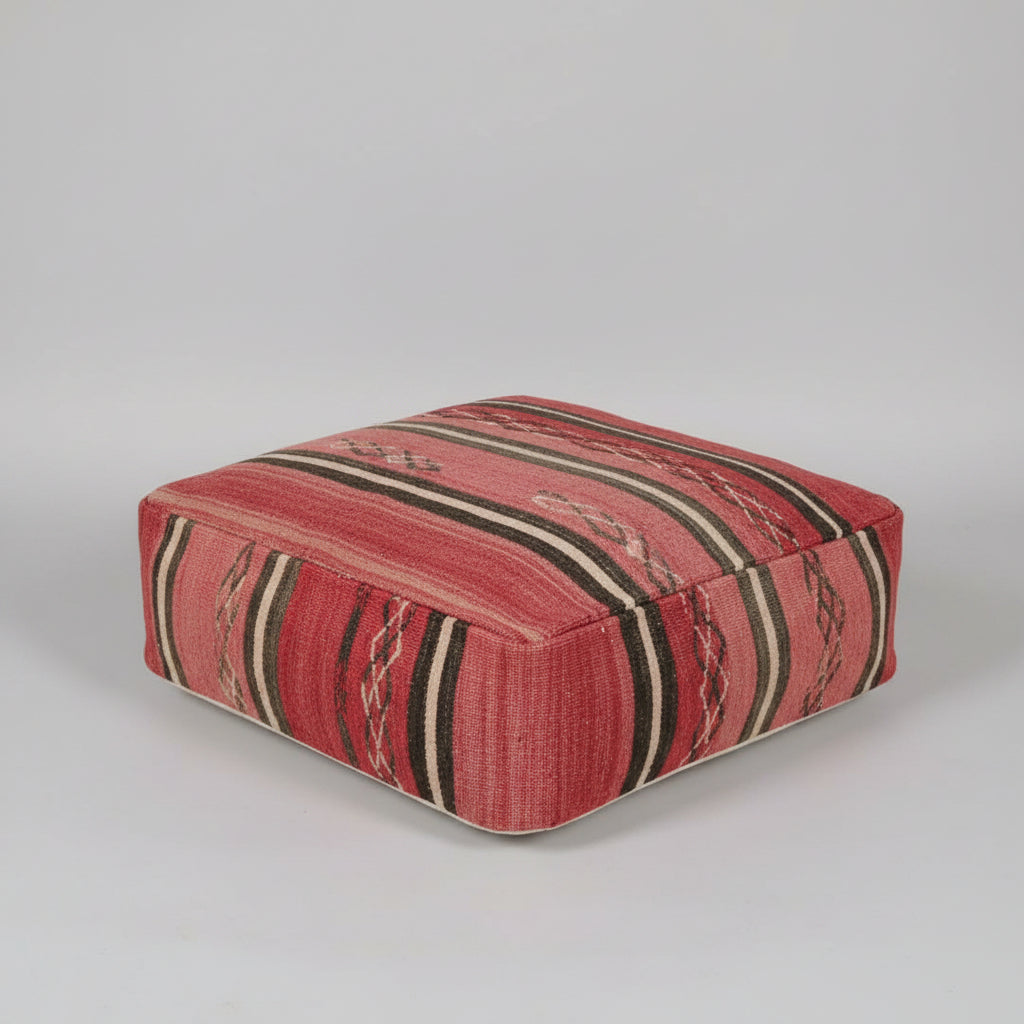 A square upholstered ottoman or pouf with a woven kilim-style pattern in red, beige, and black, photographed on a neutral light gray background.